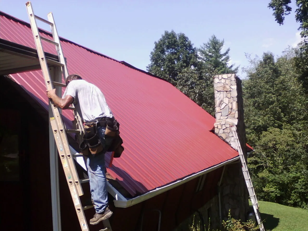 Roofer on a ladder beside a red metal roof and stone chimney — Soffit & Fascia Repair by Roofing Brothers in Addyston